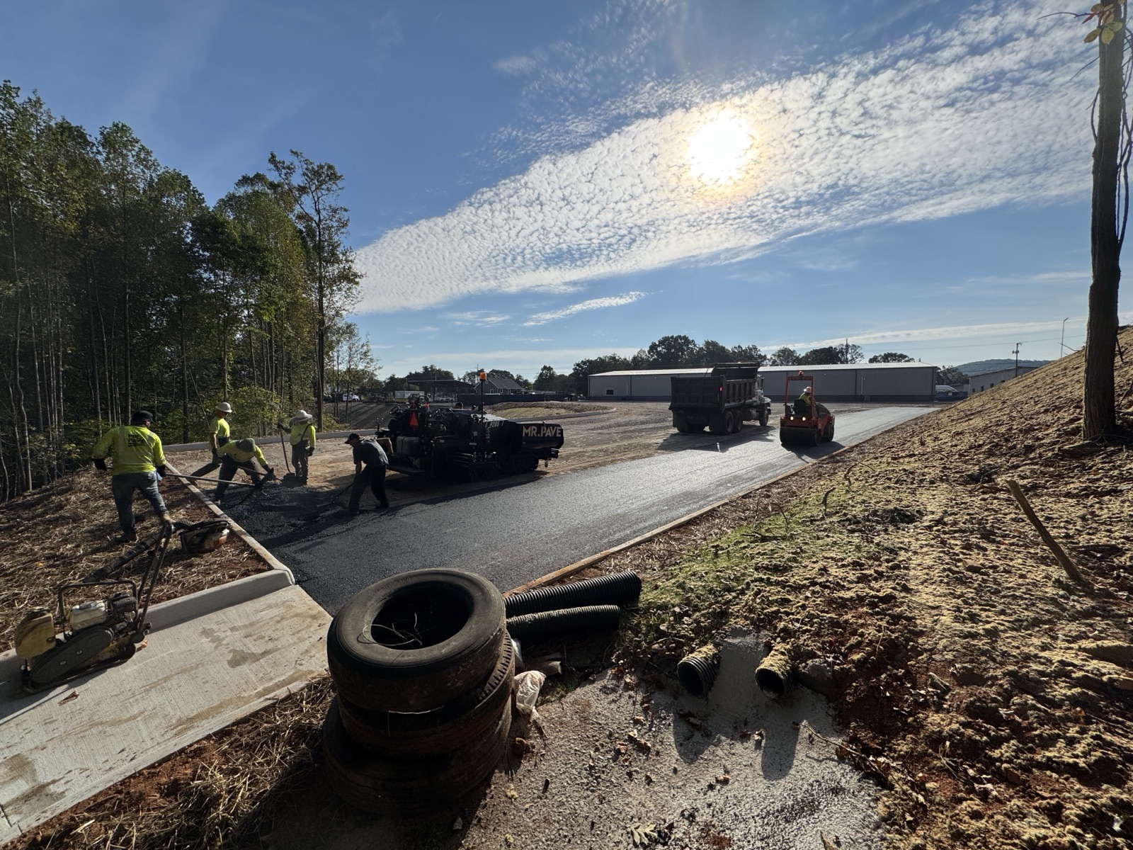 Cuadrilla de Mr. Pave pavimentando una vía con pavimentadora, camión de volteo y rodillo en el sitio